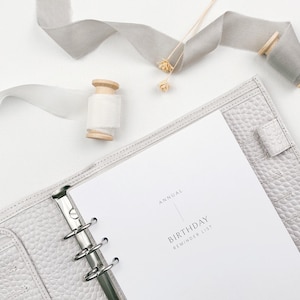 May include: A pale grey leather planner with a silver binder, open to a page that reads "Annual Birthday Reminder List." Decorative ribbon and wooden spools are arranged around the planner on a white surface.