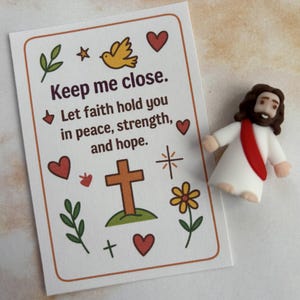 May include: A card with the text "Keep me close. Let faith hold you in peace, strength, and hope." is next to a small figurine of Jesus wearing a white robe and a red sash. The card has a cross, hearts, and other decorative elements.