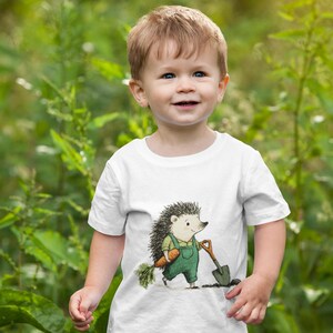 May include: A white t-shirt featuring a cartoon hedgehog in green overalls, holding a carrot and a shovel. The child is wearing blue jeans and is smiling. The background is a blurred green garden.