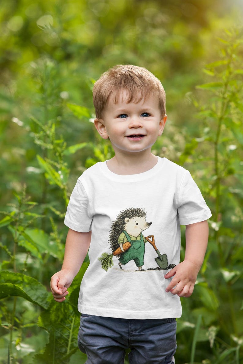 May include: A white t-shirt featuring a cartoon hedgehog in green overalls, holding a carrot and a shovel. The child is wearing blue jeans and is smiling. The background is a blurred green garden.