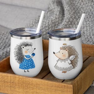 May include: Two white insulated wine tumblers with lids and straws. Each tumbler features a cartoon hedgehog illustration. One hedgehog wears a blue dress and holds a berry. The other is an angel hedgehog with a halo and wings. The tumblers sit in a wooden tray.