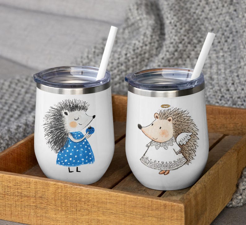 May include: Two white insulated wine tumblers with lids and straws. Each tumbler features a cartoon hedgehog illustration. One hedgehog wears a blue dress and holds a berry. The other is an angel hedgehog with a halo and wings. The tumblers sit in a wooden tray.