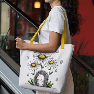 May include: White tote bag with a whimsical illustration of a hedgehog under daisies, with raindrops. The bag has yellow straps and is carried by a person wearing a white t-shirt and a pleated skirt.