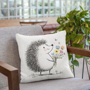 May include: White throw pillow featuring a whimsical illustration of a hedgehog holding a bouquet of colorful flowers. The hedgehog is drawn in black and white, with a smiling face and a few small butterflies. The pillow is on a gray chair.