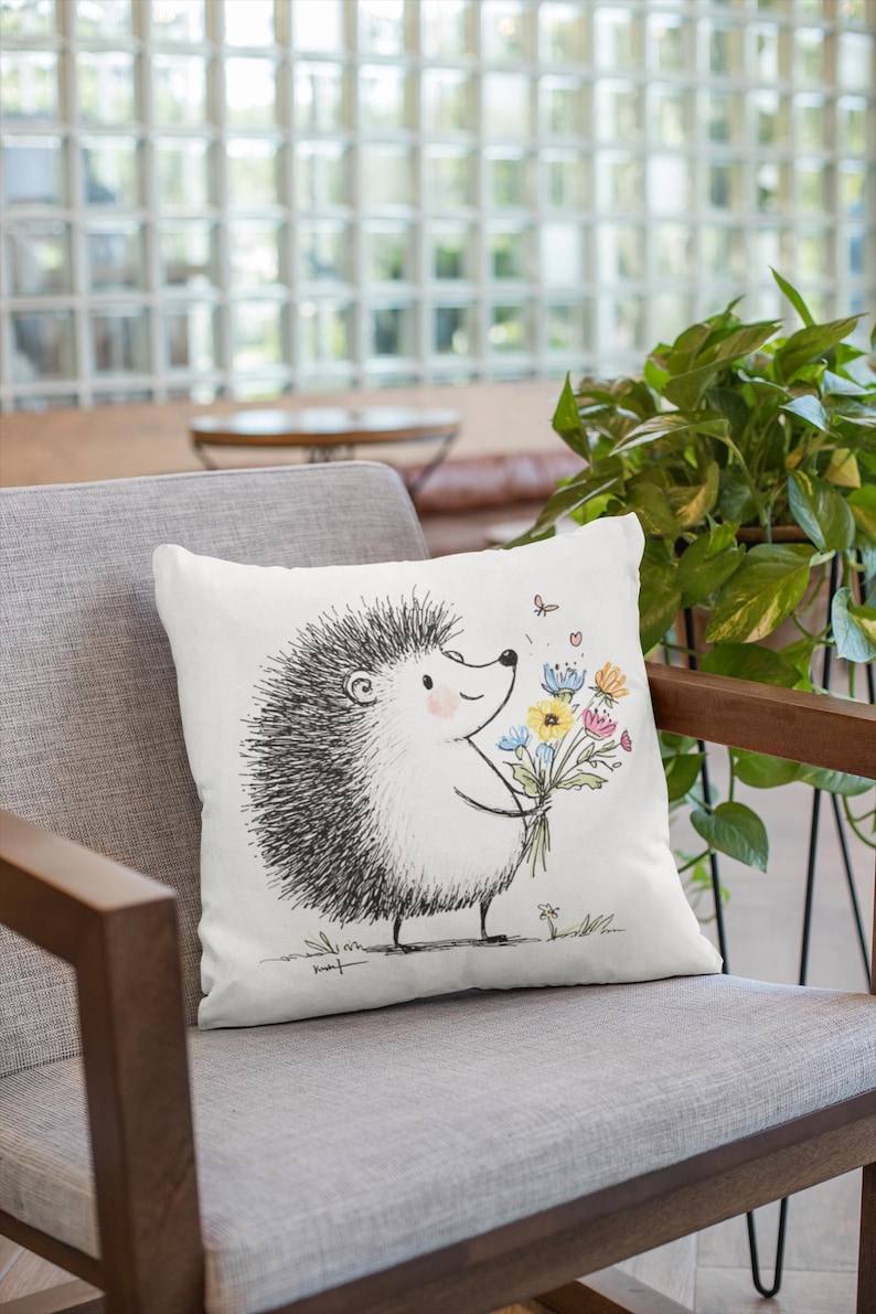 May include: White throw pillow featuring a whimsical illustration of a hedgehog holding a bouquet of colorful flowers. The hedgehog is drawn in black and white, with a smiling face and a few small butterflies. The pillow is on a gray chair.