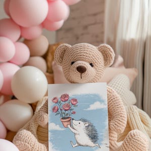 May include: A crocheted teddy bear holds a card with a drawing of a hedgehog carrying a pot of roses. The bear is light brown, and the card has a blue sky background. Pink balloons are in the background.