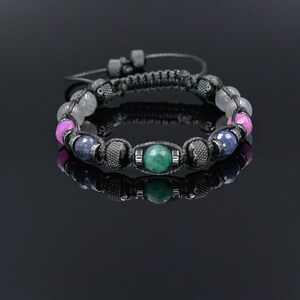 May include: A beaded bracelet featuring a central green stone, surrounded by pink, blue, and gray beads. The bracelet has a black cord and adjustable closure. The jewelry is displayed on a reflective black surface.