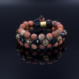 Könnte beinhalten: Ein Armband mit doppelter Schnur aus roten, braunen und schwarzen Perlen. Das Armband hat eine schwarze Schnur mit einem goldenen Verschluss.