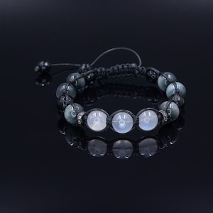 Puede incluir: Un brazalete de cordón negro con cuentas grises y blancas. El brazalete tiene un cierre de nudo.