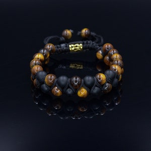 May include: A beaded bracelet featuring alternating rows of tiger eye and black onyx stones. The bracelet is strung on a black cord with a gold-colored clasp. The tiger eye beads have a warm, golden-brown color.