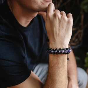 May include: A double-strand bracelet with black and purple beads. The bracelet is worn on a person's wrist. The beads are round and faceted, with a mix of black and purple colours. The bracelet is made with a black cord.