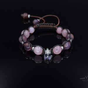 May include: A pink and purple gemstone bracelet with a silver butterfly charm. The bracelet is made with a black cord and features a silver bead with a clear stone.