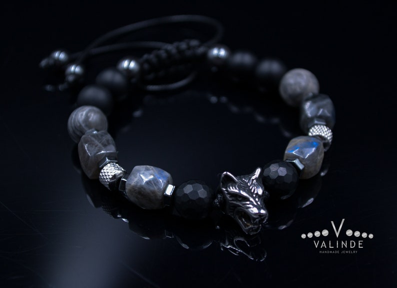 May include: A black beaded bracelet with a silver wolf charm. The beads are made of black onyx, labradorite, and hematite. The bracelet is tied with a black cord.