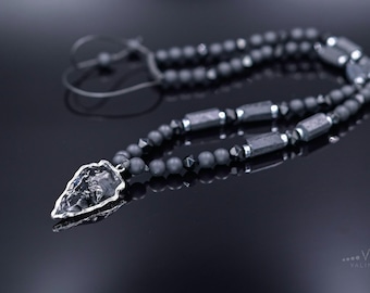 May include: A black onyx and hematite beaded necklace with a silver arrowhead pendant. The necklace is on a black surface.