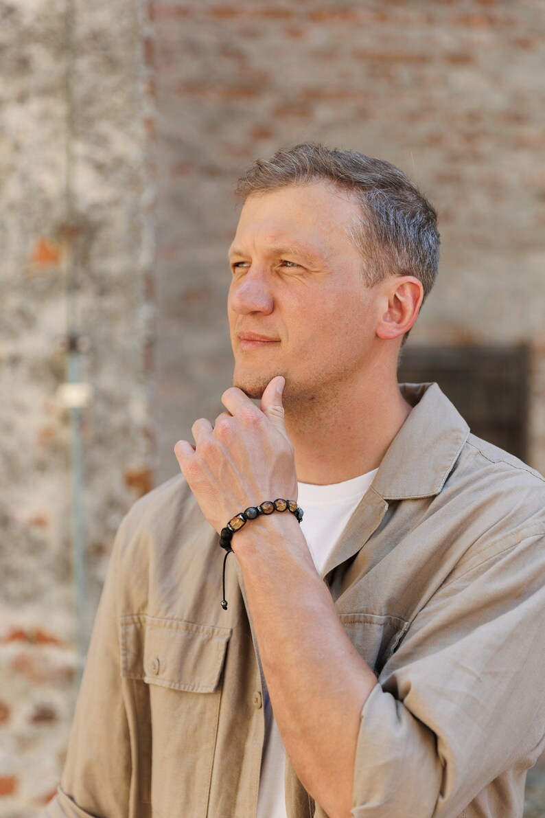Peut inclure: Un homme portant une chemise marron clair avec un bracelet en perles noires au poignet. Il regarde de c&ocirc;t&eacute; et a la main sur le menton.