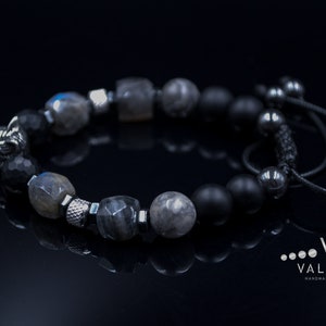 May include: A black beaded bracelet with a wolf head charm, grey and black stones, and a black cord closure. The bracelet is on a black surface.