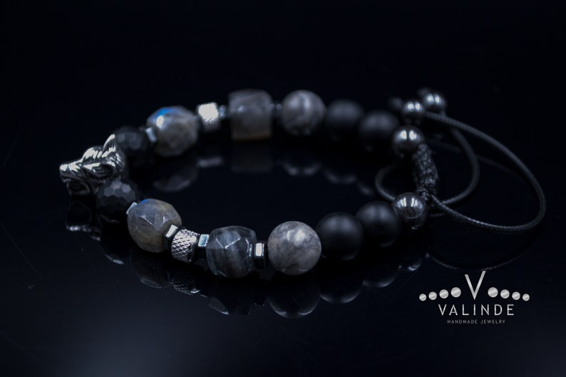 May include: A black beaded bracelet with a wolf head charm, grey and black stones, and a black cord closure. The bracelet is on a black surface.