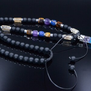 May include: A black beaded necklace with colorful gemstone accents and a clear crystal pendant. The necklace features a variety of gemstones in shades of yellow, purple, blue, and brown. The pendant is a clear crystal with colorful inclusions.