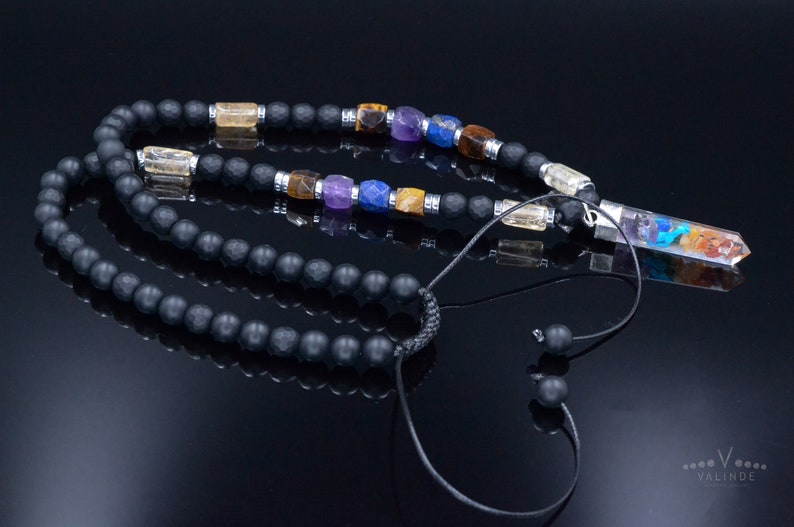 May include: A black beaded necklace with colorful gemstone accents and a clear crystal pendant. The necklace features a variety of gemstones in shades of yellow, purple, blue, and brown. The pendant is a clear crystal with colorful inclusions.