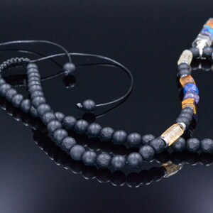 May include: A black beaded necklace with a variety of colorful gemstones and a black cord. The necklace features a mix of matte black beads, yellow, blue, and purple stones, and clear crystal accents. The necklace is displayed on a reflective black surface.