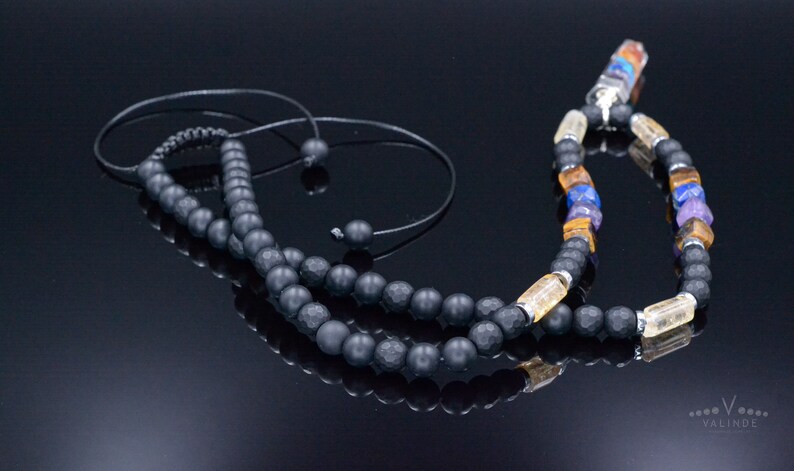 May include: A black beaded necklace with a variety of colorful gemstones and a black cord. The necklace features a mix of matte black beads, yellow, blue, and purple stones, and clear crystal accents. The necklace is displayed on a reflective black surface.