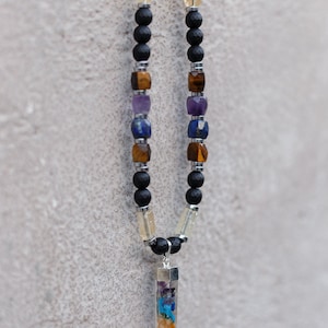 May include: A beaded necklace featuring black lava rock beads, interspersed with colorful gemstone beads in shades of yellow, brown, blue, and purple. A clear, prism-shaped pendant with layered, colorful stones hangs from the necklace.