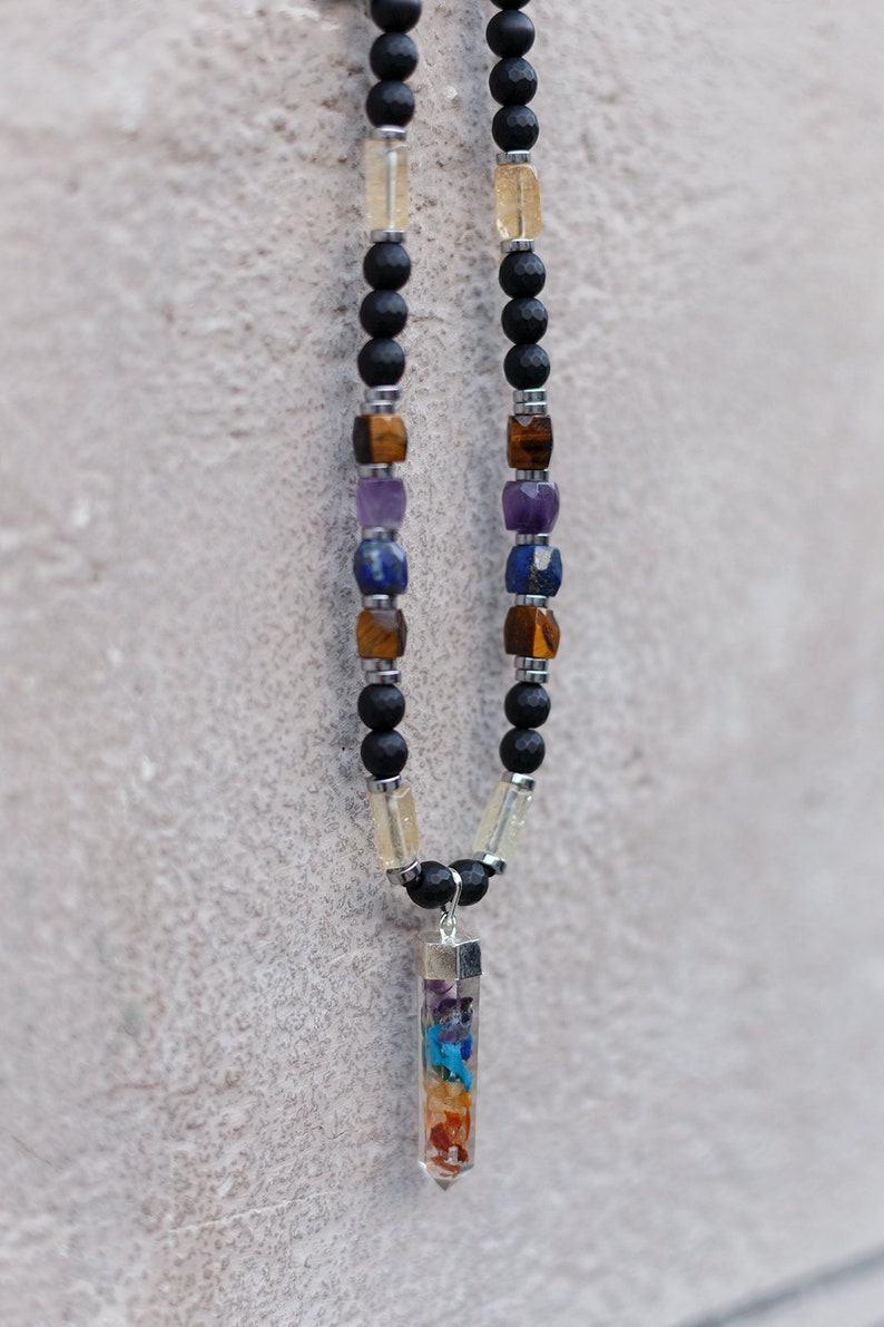May include: A beaded necklace featuring black lava rock beads, interspersed with colorful gemstone beads in shades of yellow, brown, blue, and purple. A clear, prism-shaped pendant with layered, colorful stones hangs from the necklace.