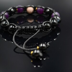 May include: A black braided bracelet with purple and black beads, a gold bead, and a black cord with a sliding knot.
