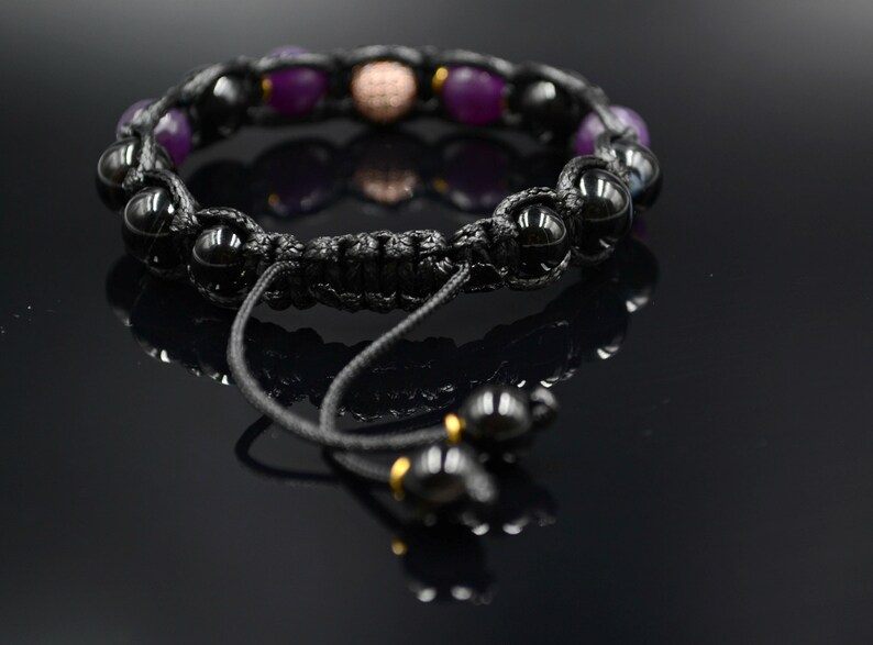 May include: A black braided bracelet with purple and black beads, a gold bead, and a black cord with a sliding knot.