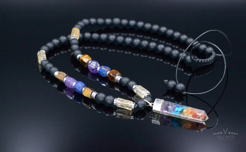 May include: A black beaded necklace with colorful gemstone accents. The necklace features a variety of stones in shades of purple, blue, yellow, and orange, and a clear rectangular pendant with a rainbow design. The necklace is displayed on a reflective black surface.