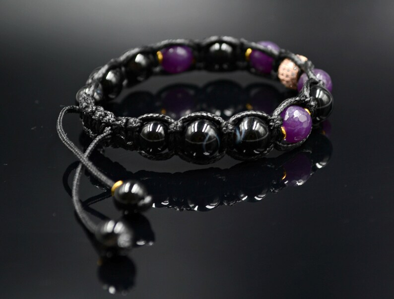 May include: Black and purple beaded bracelet with a black cord and gold accents. The bracelet has a sliding knot closure.