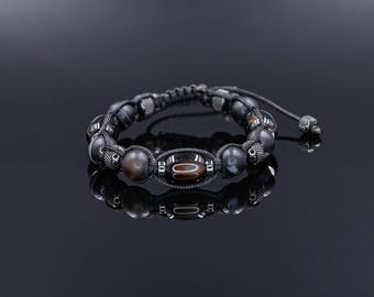 May include: A beaded bracelet featuring dark gray and brown stones, with a central oval-shaped bead. The bracelet is strung on a black cord and has adjustable ends. The bracelet is displayed on a reflective black surface.