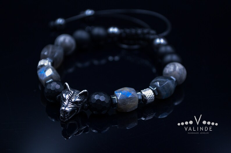 May include: A black beaded bracelet with a silver wolf charm. The beads are made of black onyx, labradorite and haematite. The bracelet is tied with a black cord.