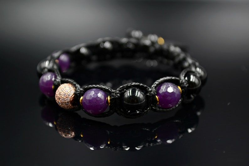 May include: A black cord bracelet with purple and black gemstone beads and a rose gold bead with clear rhinestones.