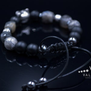 May include: A black and silver beaded bracelet with a silver charm. The bracelet has a black cord with a silver bead at the end.