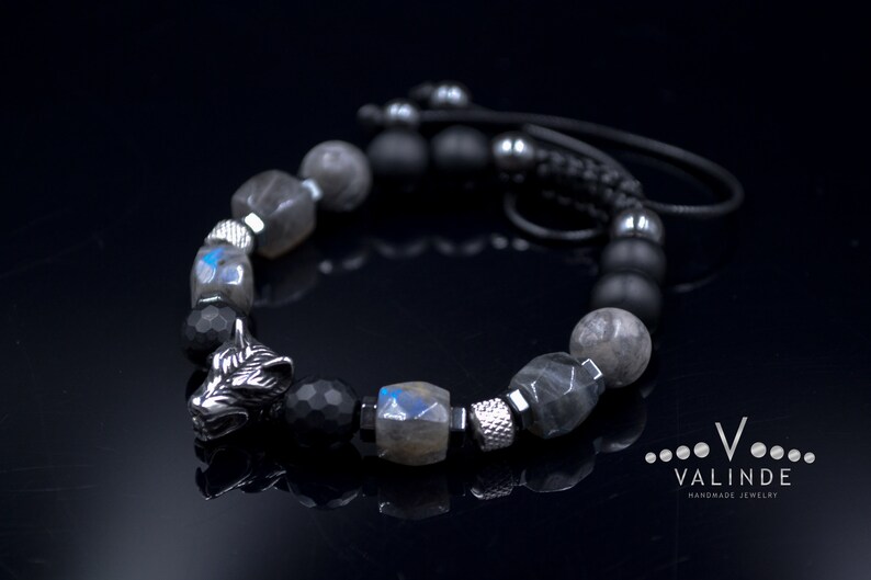 May include: A black and silver beaded bracelet with a wolf head charm. The bracelet features black onyx, labradorite and hematite beads. The wolf head charm is made of metal and has a detailed design. The bracelet is tied with a black cord.