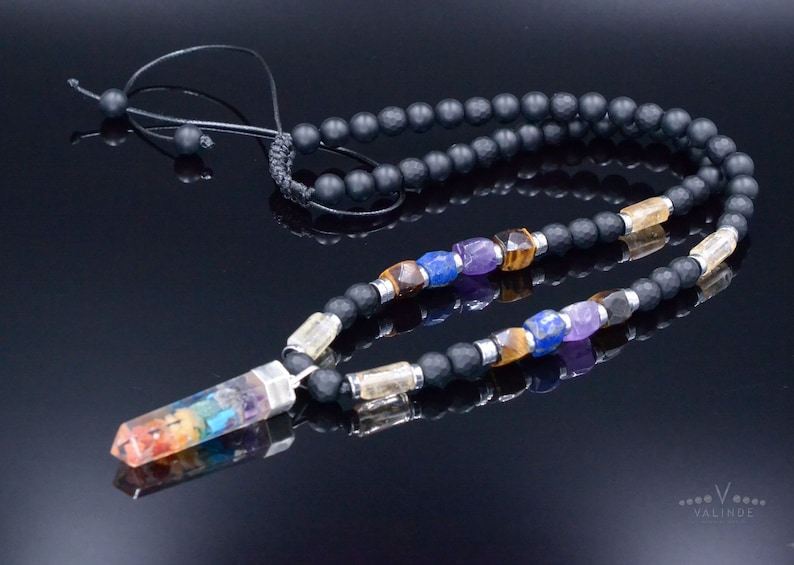 May include: A black beaded necklace with a rainbow-colored crystal pendant. The necklace features various gemstone beads in colors like purple, blue, and yellow. The pendant is a clear crystal with layers of rainbow colors. The necklace is on a reflective black surface.