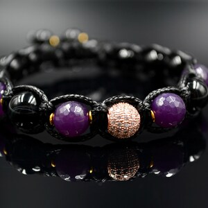 May include: A black cord bracelet with purple and rose gold beads. The bracelet has a knot design and features a round rose gold bead with clear stones.