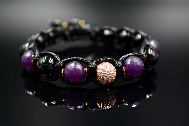 May include: A black cord bracelet with purple and rose gold beads. The bracelet has a knot design and features a round rose gold bead with clear stones.