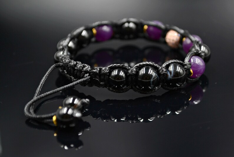 May include: Black and purple beaded bracelet with a black cord and gold accents. The bracelet has a sliding knot closure.