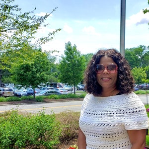 May include: A woman wearing a white crocheted top with a black skirt and a leopard print scarf. She is wearing pink sunglasses and has curly hair.
