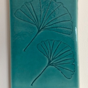 Peut inclure: Carreau en céramique bleu-vert avec deux feuilles de ginkgo en relief. Le carreau a une bordure blanche et les initiales "LG" sont écrites en bas à droite.