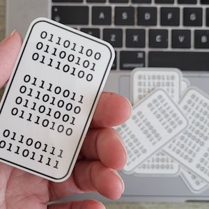 May include: White rectangular stickers featuring binary code in black. One sticker is held in a hand, with others on a laptop keyboard. The stickers have rounded corners and a white border.