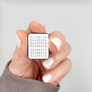 May include: A white rectangular pin with a black border, displaying binary code in black text. The pin is held between fingers against a plain white backdrop. The binary code is arranged in rows.