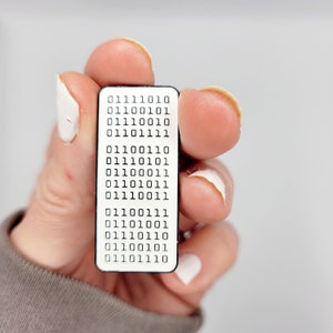 May include: A white enamel pin with a black border featuring a grid of binary code. The code reads "01110010 01100101 01110010 01101111 01110011 01110101 01100011 01110100 01110011 01110101 01100011 01110100 01110011 01110101 01100011 01110100 01110011 01110101 01100011 01110100 01110011 01110101 01100011 01110100".