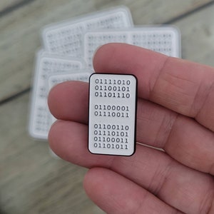 May include: A black enamel pin with a white background and a series of binary code numbers: 01111010, 01100101, 01101110, 01100001, 01110011, 01100110, 01110101, 01100011, 01101011.