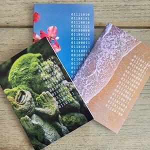May include: Three different notebooks featuring a variety of nature scenes on their covers. The first notebook has a green mossy sculpture with binary code on the cover. The second notebook has a pink flower with a blue sky and binary code on the cover. The third notebook has a beach scene with waves and sand with binary code on the cover.