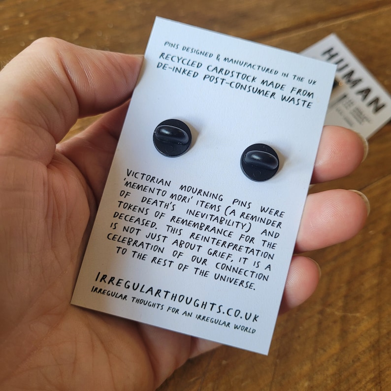 Op de afbeelding: Twee zwarte emaille-pins op een witte achtergrond. De pins zijn rond en hebben een witte rand. De tekst op de achtergrond luidt "Victorian Mourning Pins were 'Memento Mori' items (a reminder of death's inevitability) and tokens of remembrance for the deceased. This reinterpretation is not just about grief, it is a celebration of our connection to the rest of the universe." De tekst onderaan luidt "IrregularThoughts.co.uk Irregular Thoughts for an Irregular World."