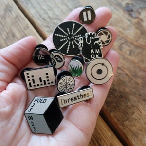Peut inclure: Une collection de broches en émail présentant divers motifs, dont un cœur avec le texte "I AM OK", une cible avec le texte "STRATEGIC", un cercle avec un graphique linéaire, un cercle avec le texte "BE", un cercle avec une cible, un cercle avec un graphique linéaire, un cercle avec le texte "HOLD 2 3 IN 2 3", un cercle avec le texte "[breathes]", un cercle avec un graphique linéaire, un cercle avec le texte "478", un cercle avec un symbole de pause, un cercle avec un objectif d'appareil photo et un cercle avec un motif d'herbe verte.