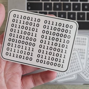 May include: A white sticker with a black border and a grid of binary code. The code is arranged in two columns, with each column containing eight rows of binary numbers. The sticker is being held in a hand.